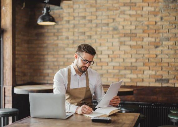 Restaurantejer med forklæde gennemgår dokumenter og ubetalte fakturaer ved en bærbar computer.